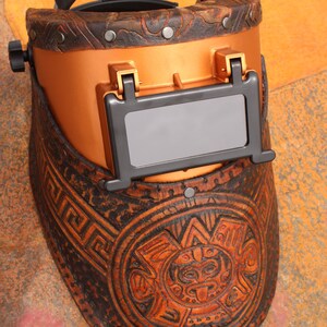 May include: A brown leather welding helmet with a copper-coloured face shield and intricate carvings. The helmet has a black leather strap and a dark grey viewing window.