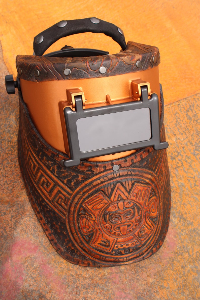 May include: A brown leather welding helmet with a copper-coloured face shield and intricate carvings. The helmet has a black leather strap and a dark grey viewing window.