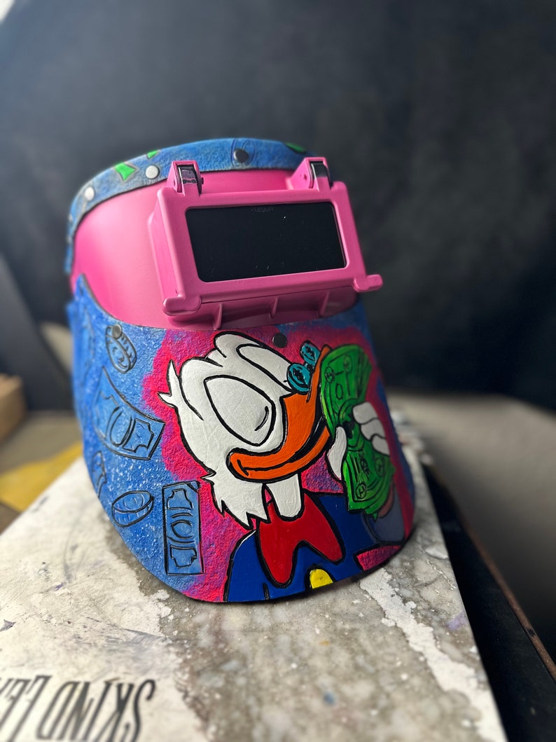May include: A pink welding helmet with a cartoon illustration of a duck wearing a blue shirt and red bow tie, holding a stack of money. The helmet has a clear viewing window and is decorated with a blue and white pattern of dollar bills.