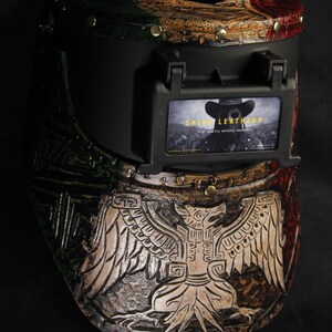 Op de afbeelding: Een op maat gemaakte lashelm met een gedetailleerd, gebeeldhouwd ontwerp. De helm heeft een zwart vizier en een decoratief patroon met rode, groene en bruine accenten. Het ontwerp bevat een adelaarsmotief en de tekst "SKIN LEATHERS".