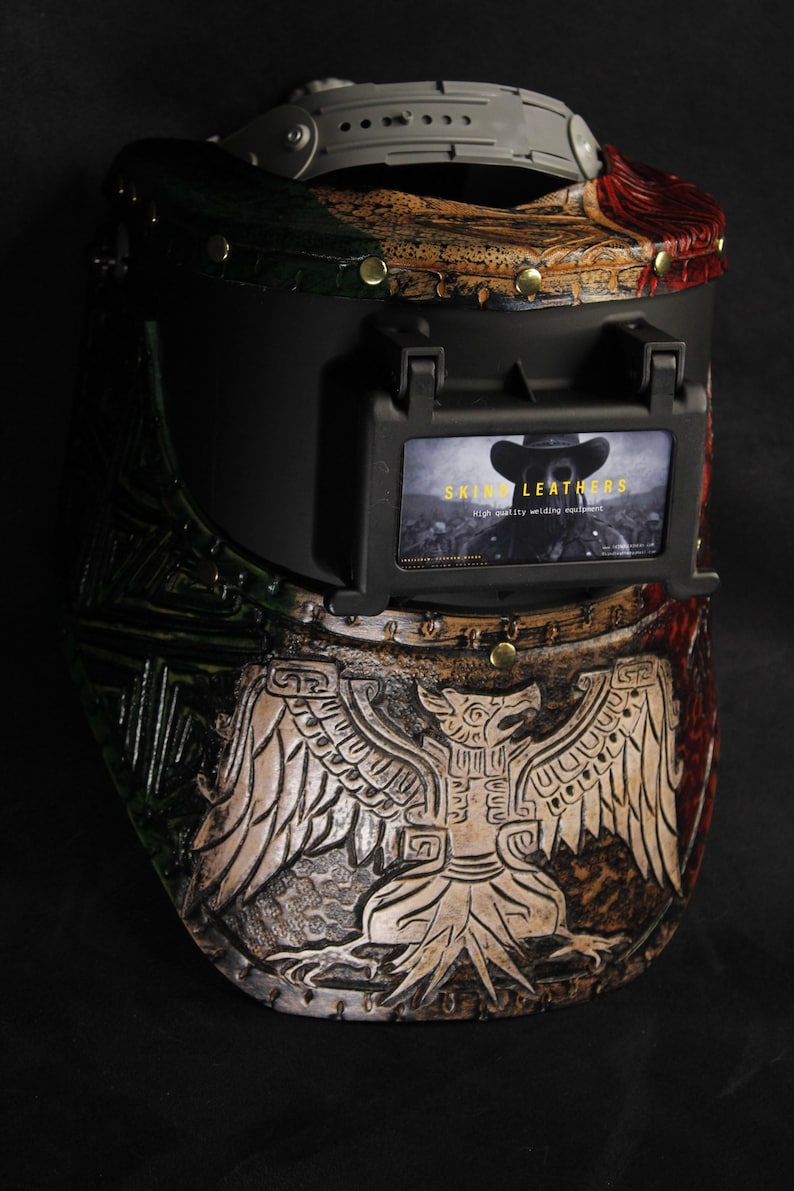 Op de afbeelding: Een op maat gemaakte lashelm met een gedetailleerd, gebeeldhouwd ontwerp. De helm heeft een zwart vizier en een decoratief patroon met rode, groene en bruine accenten. Het ontwerp bevat een adelaarsmotief en de tekst "SKIN LEATHERS".