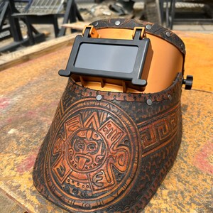 May include: A brown leather welding helmet with a gold-coloured metal frame and a black viewing window. The helmet has an intricate design of a circular pattern with a face in the centre.