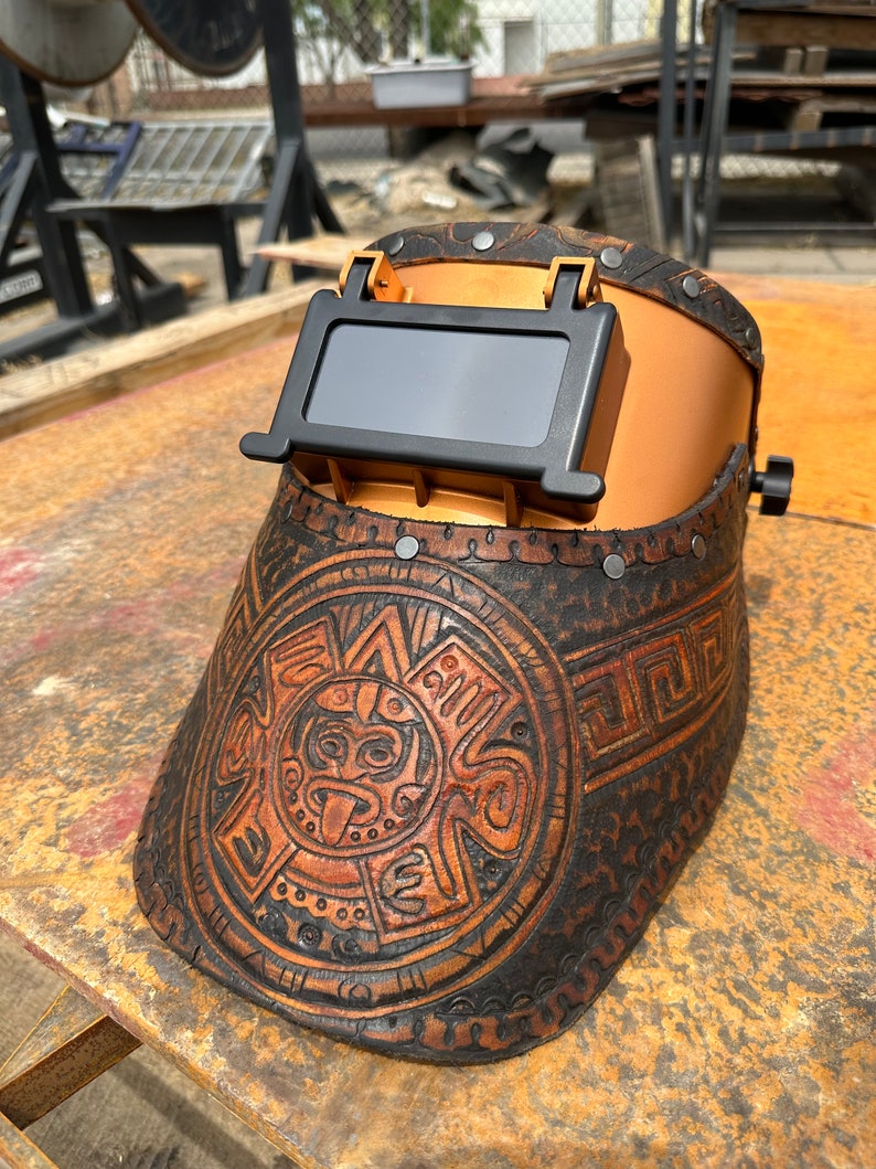 May include: A brown leather welding helmet with a gold-coloured metal frame and a black viewing window. The helmet has an intricate design of a circular pattern with a face in the centre.