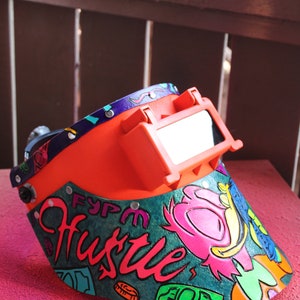 May include: A bright orange welding helmet with a clear visor. The helmet is decorated with a colorful design featuring the word "Hustle" in a stylized font, along with other graphic elements.