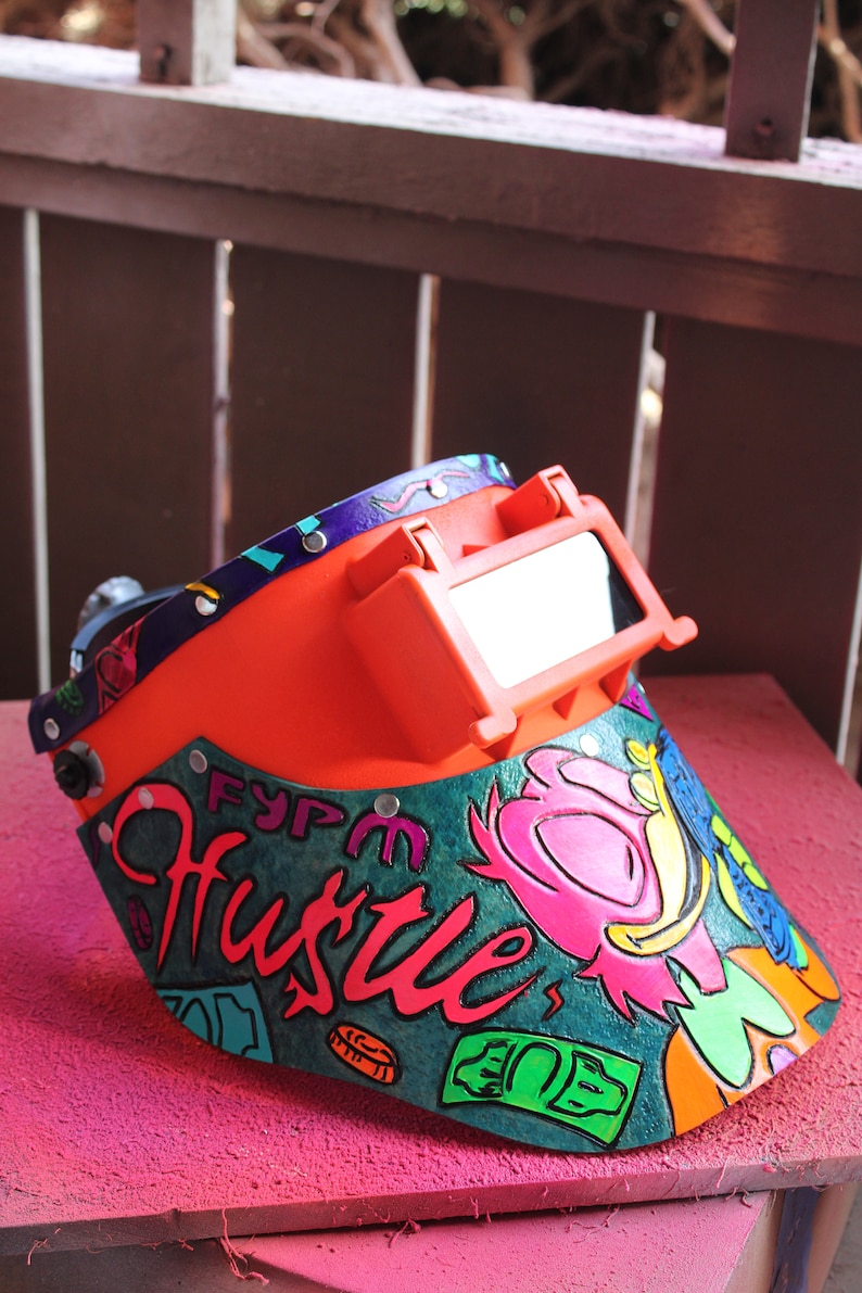 May include: A bright orange welding helmet with a clear visor. The helmet is decorated with a colorful design featuring the word "Hustle" in a stylized font, along with other graphic elements.