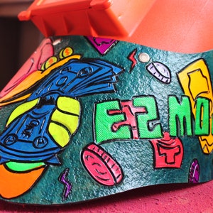 May include: A green, orange, blue, and yellow painted welding helmet with the words "EZ MONEY" in green letters. The helmet is decorated with a cartoon image of money and coins.