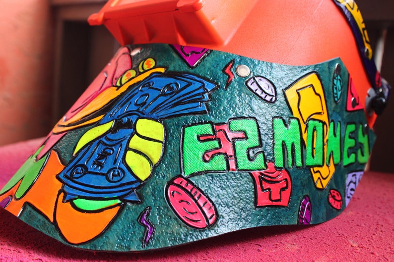 May include: A green, orange, blue, and yellow painted welding helmet with the words "EZ MONEY" in green letters. The helmet is decorated with a cartoon image of money and coins.