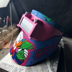 May include: A pink welding helmet with a blue and green design. The helmet has a cartoon image of a duck holding money and the word "HUSTLE" written on it.