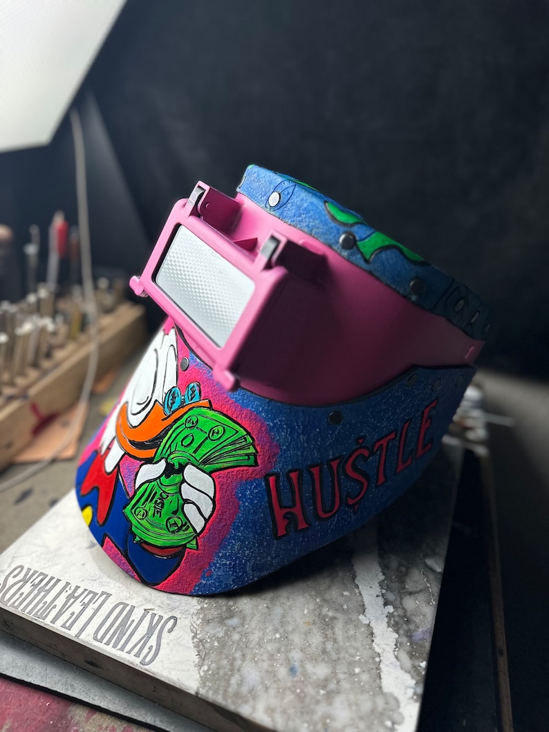 May include: A pink welding helmet with a blue and green design. The helmet has a cartoon image of a duck holding money and the word "HUSTLE" written on it.