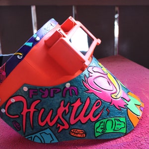 May include: A bright orange welding helmet with a clear visor. The helmet is decorated with a colorful design featuring the words "FYPM Hustle" and a cartoon character. The design also includes dollar bills and coins.