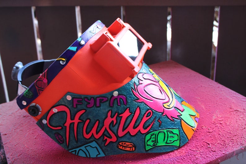May include: A bright orange welding helmet with a clear visor. The helmet is decorated with a colorful design featuring the words "FYPM Hustle" and a cartoon character. The design also includes dollar bills and coins.