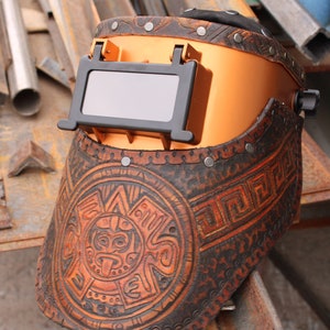 May include: A brown leather welding helmet with a copper-coloured face shield and a decorative Aztec-inspired design. The helmet has a black viewing window and is adorned with metal rivets.