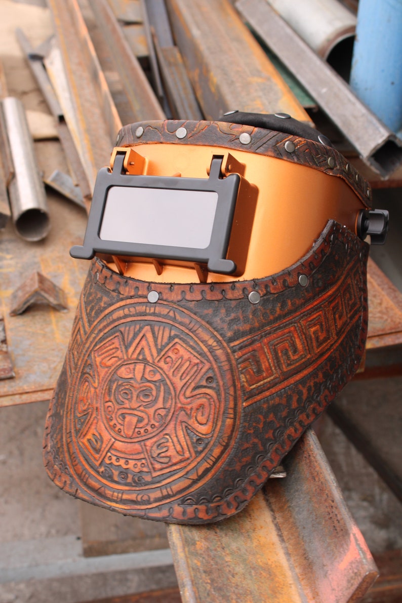 May include: A brown leather welding helmet with a copper-coloured face shield and a decorative Aztec-inspired design. The helmet has a black viewing window and is adorned with metal rivets.