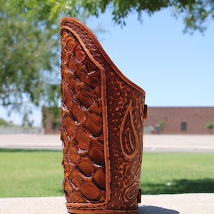 May include: A brown leather arm guard with a decorative pattern. The guard is made of thick leather and has a textured surface.