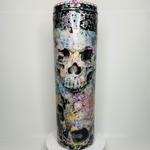 May include: A tall, cylindrical tumbler with a skull and floral design. The tumbler features a black and white skull graphic with colorful floral accents. The top is black with iridescent details and a beaded rim. A pink straw is inserted into the lid.