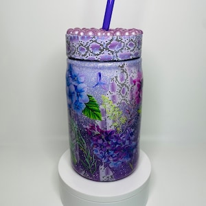 May include: Purple and green floral patterned tumbler with a glittery finish and a purple lid with pearl accents. The tumbler has a straw.