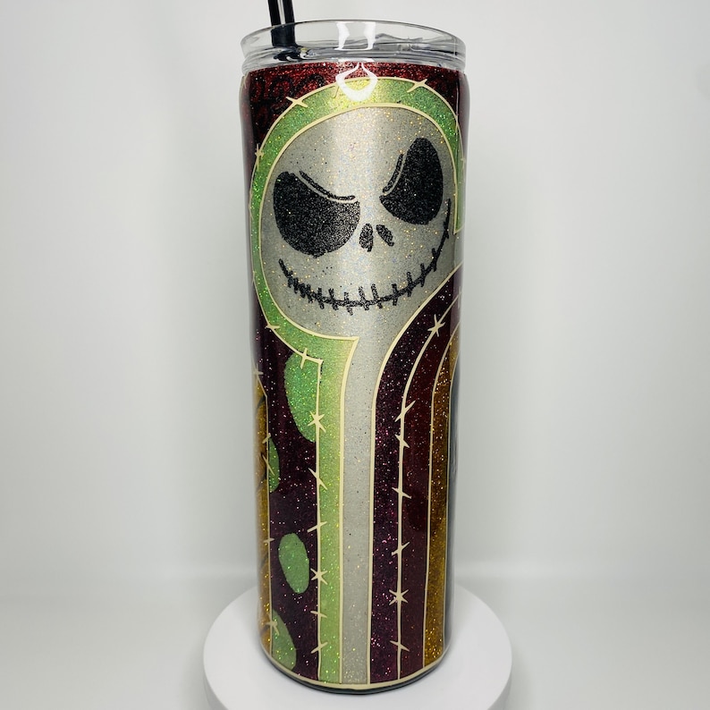 Op de afbeelding: Een roestvrijstalen tumbler met een glinsterende afwerking. De tumbler heeft een gestreept ontwerp in groen, goud en bordeauxrood met een wit silhouet van Jack Skellingtons gezicht uit de film The Nightmare Before Christmas.