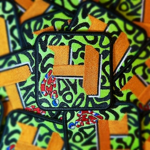 Puede incluir: Parche cuadrado bordado con fondo verde neón y formas abstractas negras. El parche presenta un diseño de letra "F" naranja y un gráfico de serpiente roja, blanca y azul. Los parches están apilados.