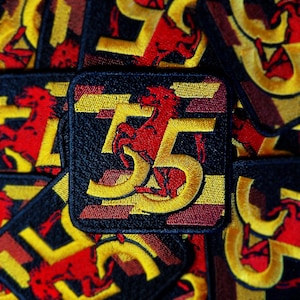 May include: Close-up of a red and yellow embroidered patch with the number "75" and a stylized horse design. The patch is surrounded by other similar patches.