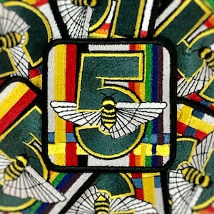 May include: Embroidered patch featuring a stylized bee with outstretched wings, set against a green background with the number "5". The patch has a black border and colorful stripes. Other similar patches are visible.