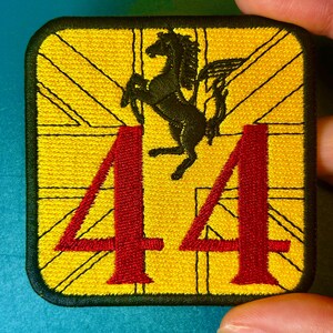 以下が含まれることがあります： 黄色い背景に黒い跳ね馬、赤い数字44、ユニオンジャックのデザインが施された刺繍パッチ。パッチには黒い縁取りがあります。