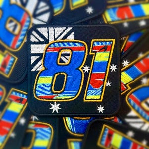 Puede incluir: Parche cuadrado negro con el número "81" bordado en azul, amarillo y rojo. Presenta un diseño de bandera blanca y negra y detalles de estrellas blancas. Los parches son probablemente para ropa o accesorios.
