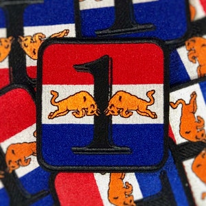 May include: Square patch with a red, white, and blue design, featuring a black number 1 and two orange bull illustrations. The patch has a black border and a textured surface, suitable for apparel.