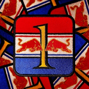 Op de afbeelding: Een geborduurde patch in rood, wit en blauw met een groot gouden nummer "1" in het midden. Twee rode stieren flankeren het nummer, met hun gezichten in tegengestelde richtingen. De patch heeft een zwarte rand.