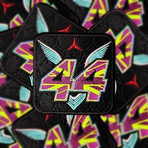 Könnte beinhalten: Ein schwarzer gestickter Patch mit der Nummer "44" in einem rosa-gelben Farbschema. Die Nummer ist von einem blauen und weißen Design mit Flügeln umgeben. Der Patch hat einen roten Stern in der Mitte.