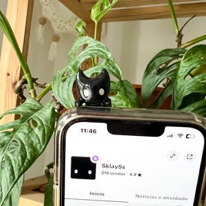 Op de afbeelding: Een zwarte telefoonhoes met een klein zwart beeldje erop. Het telefoonscherm toont een Etsy-winkel met de naam "SklaySs" en de tekst "219 vendas 4.9".