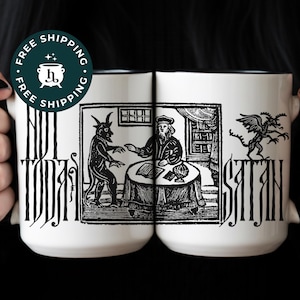 Peut inclure: Deux mugs en céramique blanche avec une illustration en noir et blanc d'un diable et d'un homme à une table. Les mugs portent les mots "NOT TODAY SATAN" en lettres gothiques noires. Les mugs sont tenus par une personne avec du vernis à ongles noir.