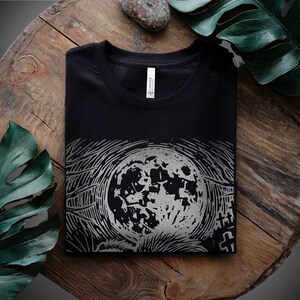 A folk punk linocut shirt with a black and white image of a crust punk opossum howling at the moon in a graveyard. Awesome opossum shirt for alt clothing folks