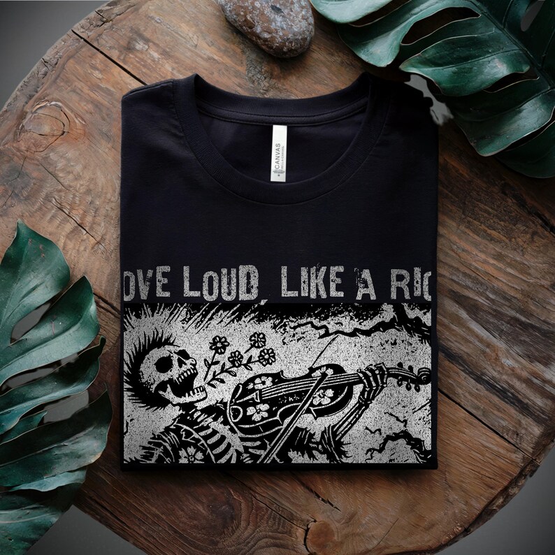 A Folk Punk Linocut shirt with a black and white image of a crust punk Skeleton playing the fiddle in a graveyard. These Offensive T-Shirts have the words Love Loud, Like A Riot; at the top.