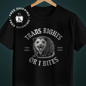 May include: Black t-shirt featuring a snarling opossum graphic with the text "TRANS RIGHTS" and "OR I BITES". The words "TRANS RIGHTS" and "HUMAN RIGHTS" are also printed on the shirt. The design is in shades of grey.