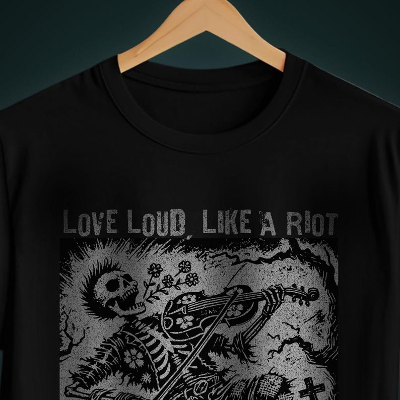 A Folk Punk Linocut shirt with a black and white image of a crust punk Skeleton playing the fiddle in a graveyard. These Offensive T-Shirts have the words Love Loud, Like A Riot; at the top.
