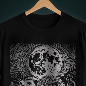 A folk punk linocut shirt with a black and white image of a crust punk opossum howling at the moon in a graveyard. Awesome opossum shirt for alt clothing folks