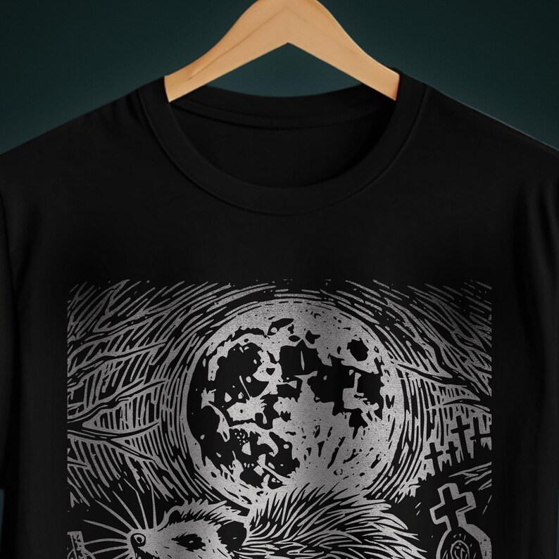 A folk punk linocut shirt with a black and white image of a crust punk opossum howling at the moon in a graveyard. Awesome opossum shirt for alt clothing folks
