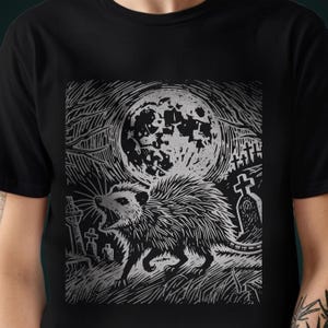 A folk punk linocut shirt with a black and white image of a crust punk opossum howling at the moon in a graveyard. Awesome opossum shirt for alt clothing folks