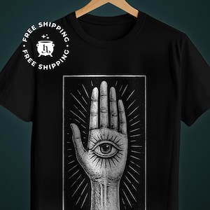 May include: Black t-shirt featuring a detailed illustration of a hand with an eye in the palm. The design is in a grayscale color scheme, with radiating lines emanating from the hand. The shirt is displayed on a wooden hanger.