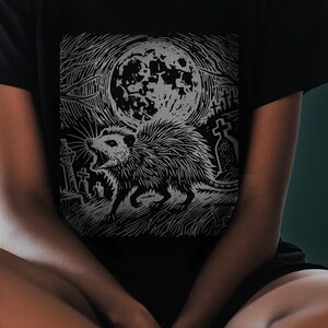 A folk punk linocut shirt with a black and white image of a crust punk opossum howling at the moon in a graveyard. Awesome opossum shirt for alt clothing folks