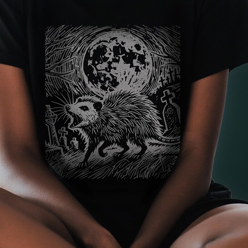 A folk punk linocut shirt with a black and white image of a crust punk opossum howling at the moon in a graveyard. Awesome opossum shirt for alt clothing folks