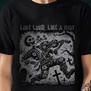 A Folk Punk Linocut shirt with a black and white image of a crust punk Skeleton playing the fiddle in a graveyard. These Offensive T-Shirts have the words Love Loud, Like A Riot; at the top.