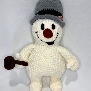 Peut inclure: Un jouet de bonhomme de neige en crochet avec un chapeau haut de forme gris, un nez rouge et une pipe brune. Le bonhomme de neige sourit et a des yeux noirs.