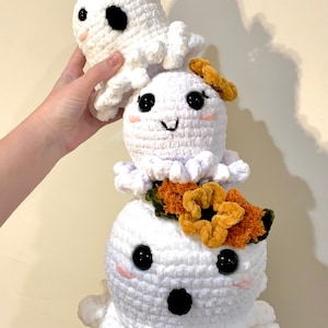 May include: A stack of three crocheted octopus plushies. The bottom octopus is white with black eyes and a surprised expression. The middle octopus is white with black eyes and a happy expression. The top octopus is white with black eyes and a witch's hat. The middle octopus has a flower on its head.