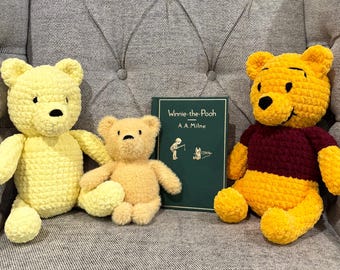 Op de afbeelding: Drie gehaakte Winnie de Poeh-beren in geel en goud, met een bordeauxrode trui op één. Een groen boek met de titel "Winnie-the-Pooh" van A.A. Milne staat tussen de beren. De beren zitten op een grijze gestoffeerde stoel.