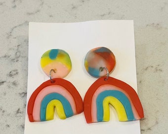 Könnte beinhalten: Ein Paar handgefertigte Regenbogen-Ohrringe. Jeder Ohrring hat einen roten Bogen mit rosa, blauen und gelben Schichten. Der obere Teil jedes Ohrrings ist ein farbenfroher, marmorierter Kreis. Die Ohrringe werden auf einer weißen Karte präsentiert.