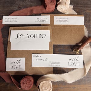 May include: A collection of wedding invitation stationery, including kraft paper envelopes, white cards, and address labels. The cards feature elegant calligraphy with phrases like "So. YOU IN?" and "with LOVE." Decorative ribbons and a wax seal add to the aesthetic.
