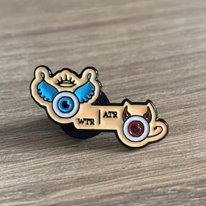 May include: A gold-colored enamel pin with a blue eye with wings on one side and a red eye with horns on the other side. The pin says "WTR | ATR" in the middle.