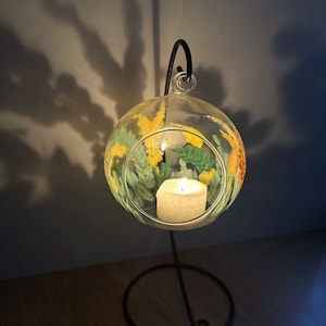 Op de afbeelding: Een glazen kaarshouder met een bloemenmotief in tinten geel en groen. De houder hangt aan een zwarte metalen standaard en heeft een brandende kaars erin.