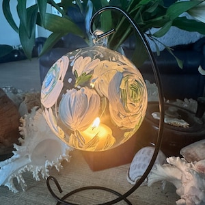 Op de afbeelding: Een heldere glazen bol met een wit bloemmotief erop geschilderd. De bol wordt van binnenuit verlicht met een kaars en hangt aan een zwarte metalen standaard.